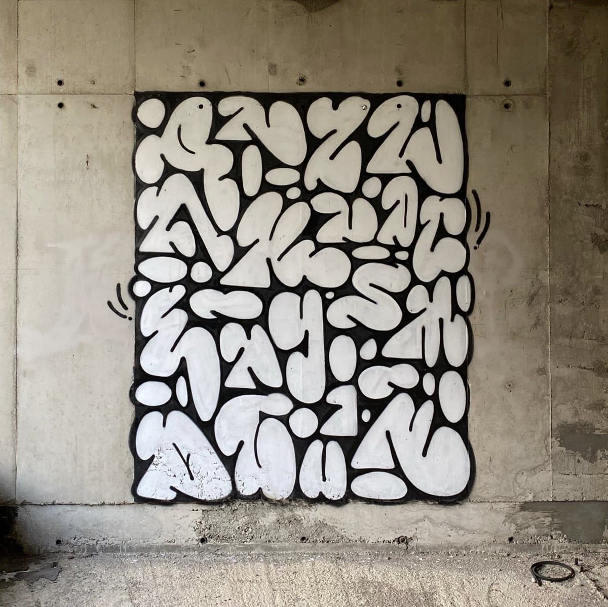 A square piece of black-and-white graffiti art is painted on a concrete wall, featuring abstract, bubble-like shapes that resemble stylized letters. The artwork is centered on the wall, which shows signs of wear and weathering, with small motion lines drawn on either side of the piece.