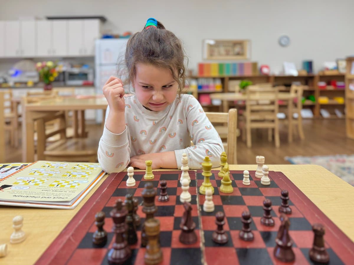 A young girl with a focused expression sits at a table playing a game of chess, with her fist clenched in a gesture of determination. A chess instruction book is open on the table next to the board, and the background shows a classroom or playroom setting with wooden furniture and shelves.