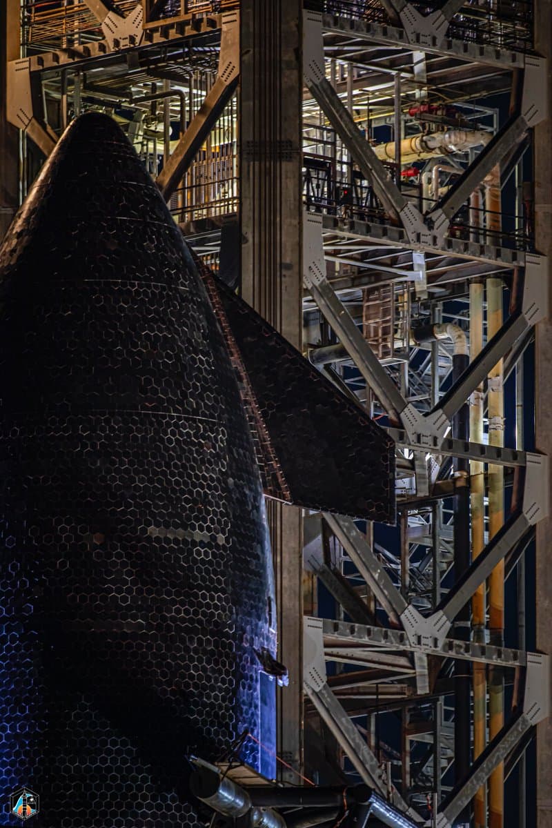 A close-up photograph of the nose and wing of a black, heat-shielded spacecraft, likely a SpaceX Starship, positioned next to a large, multi-level steel launch tower structure.