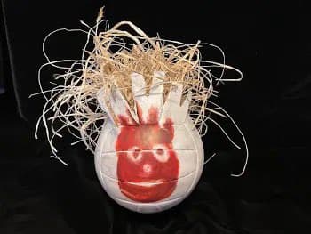A white, spherical object resembling a ball sits on a black surface, featuring a red painted face with a wide mouth and round glasses. A large, messy tuft of dried, straw-like fibers protrudes from the top of the object.