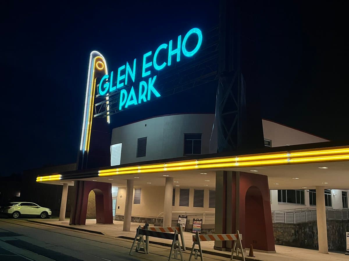A nighttime photograph of the exterior of the Glen Echo Park building, featuring a large illuminated blue neon sign and yellow marquee lights. A white car is parked on the left, and construction barriers are visible in the foreground.
