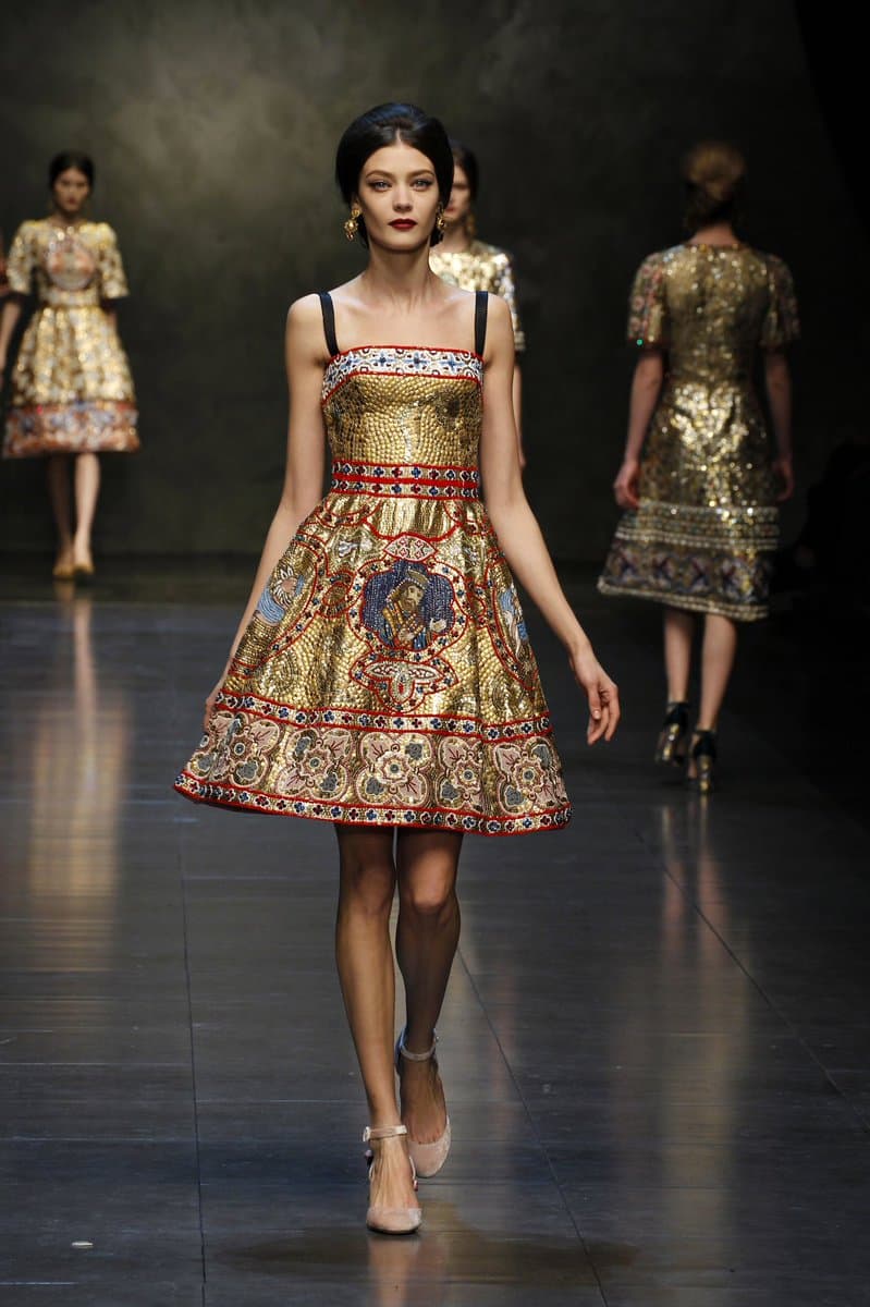 A fashion model walks a runway wearing a gold, Byzantine-style dress featuring intricate mosaic patterns and a central religious icon. Other models in similar gold garments are visible in the background.