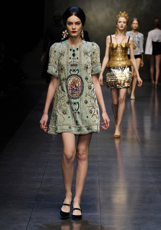 A fashion runway photograph showing a model walking down a dark, reflective catwalk wearing a pale green, heavily embroidered dress with a large central medallion. Behind her, another model walks in a gold, sequined mini-dress and a crown-like headpiece.