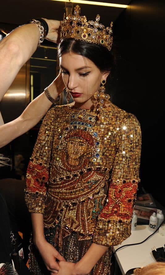 A woman wearing an ornate, gold-leafed Byzantine-style dress and a jeweled crown is being styled by a person whose hands are visible adjusting the crown.