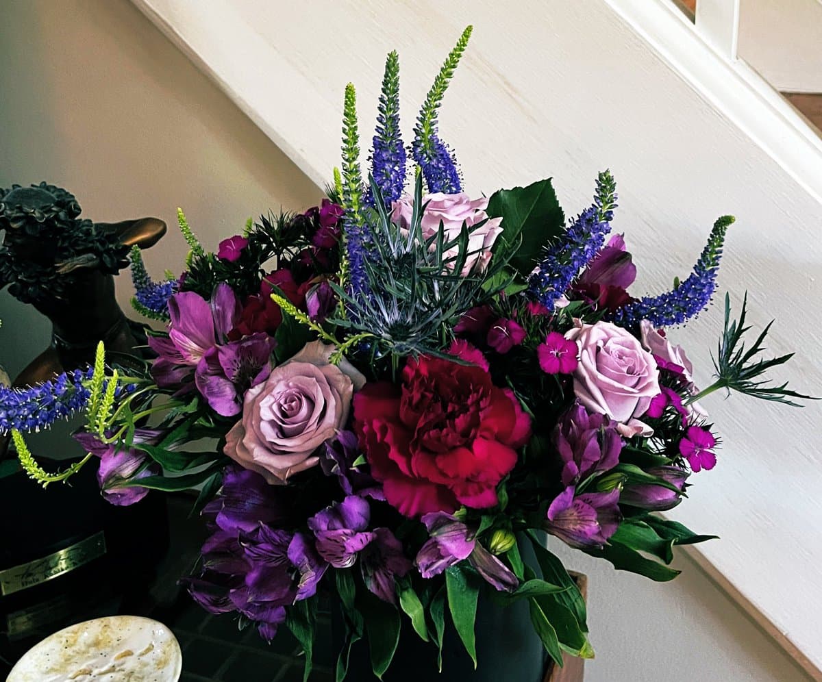 A vibrant floral arrangement featuring deep purple and pink roses, alstroemeria, and red peonies, accented with tall blue spikes and greenery, displayed in a dark vase next to a small statue.