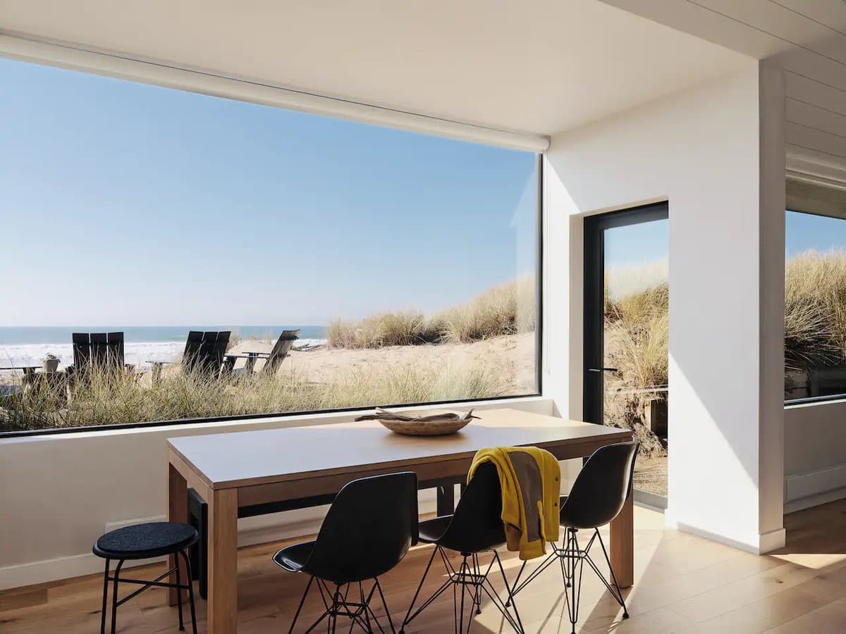 A modern dining area inside a beach house features a wooden table and black chairs positioned in front of a large window that offers a view of the ocean and sandy dunes. A yellow jacket is draped over one of the chairs, and the room is illuminated by bright natural light.