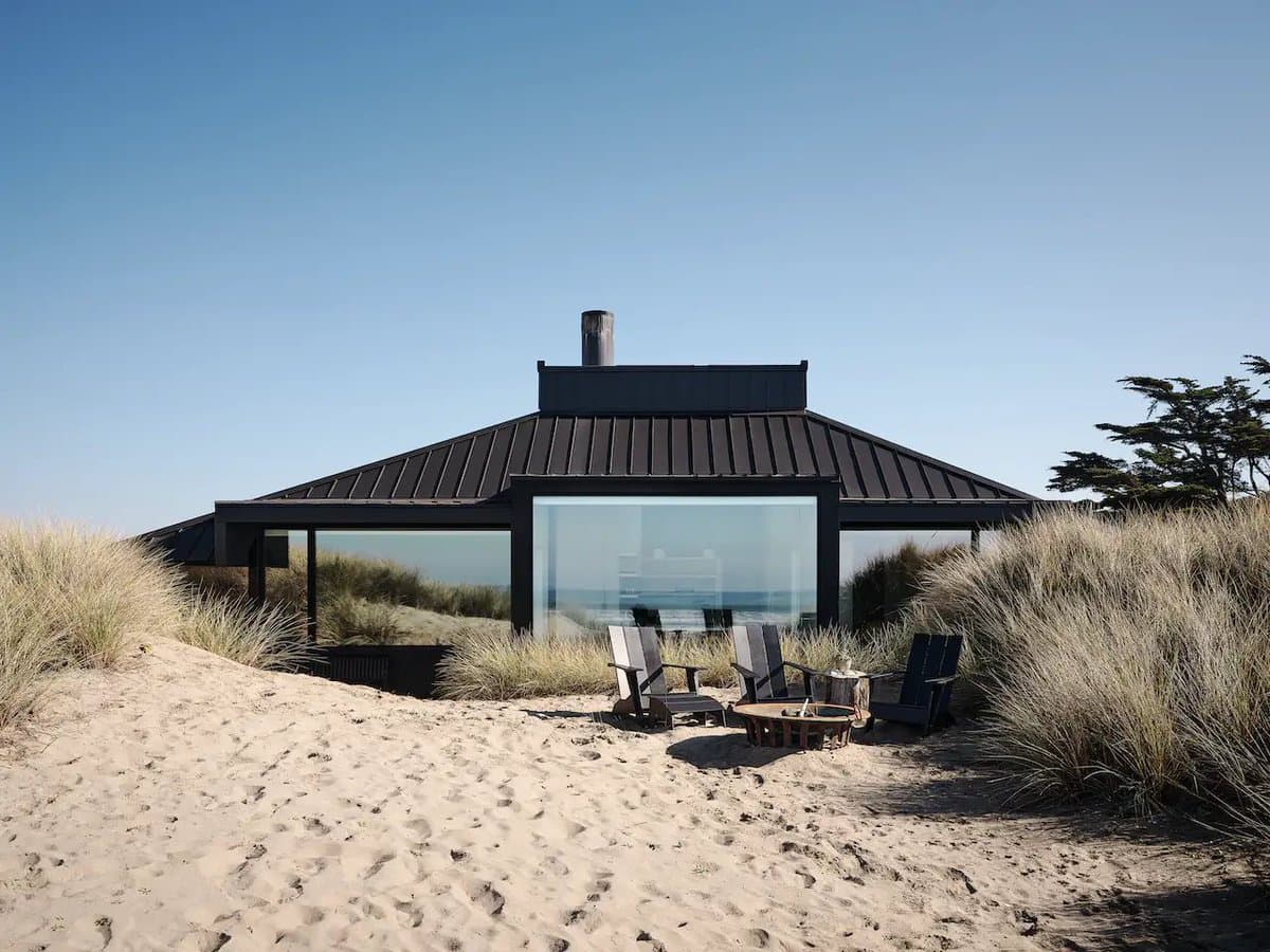 A modern, low-slung house with a dark metal roof and large glass windows sits on a sandy beach, surrounded by tall dune grass. Three outdoor chairs and a small table are arranged on the sand in front of the structure, facing the ocean.