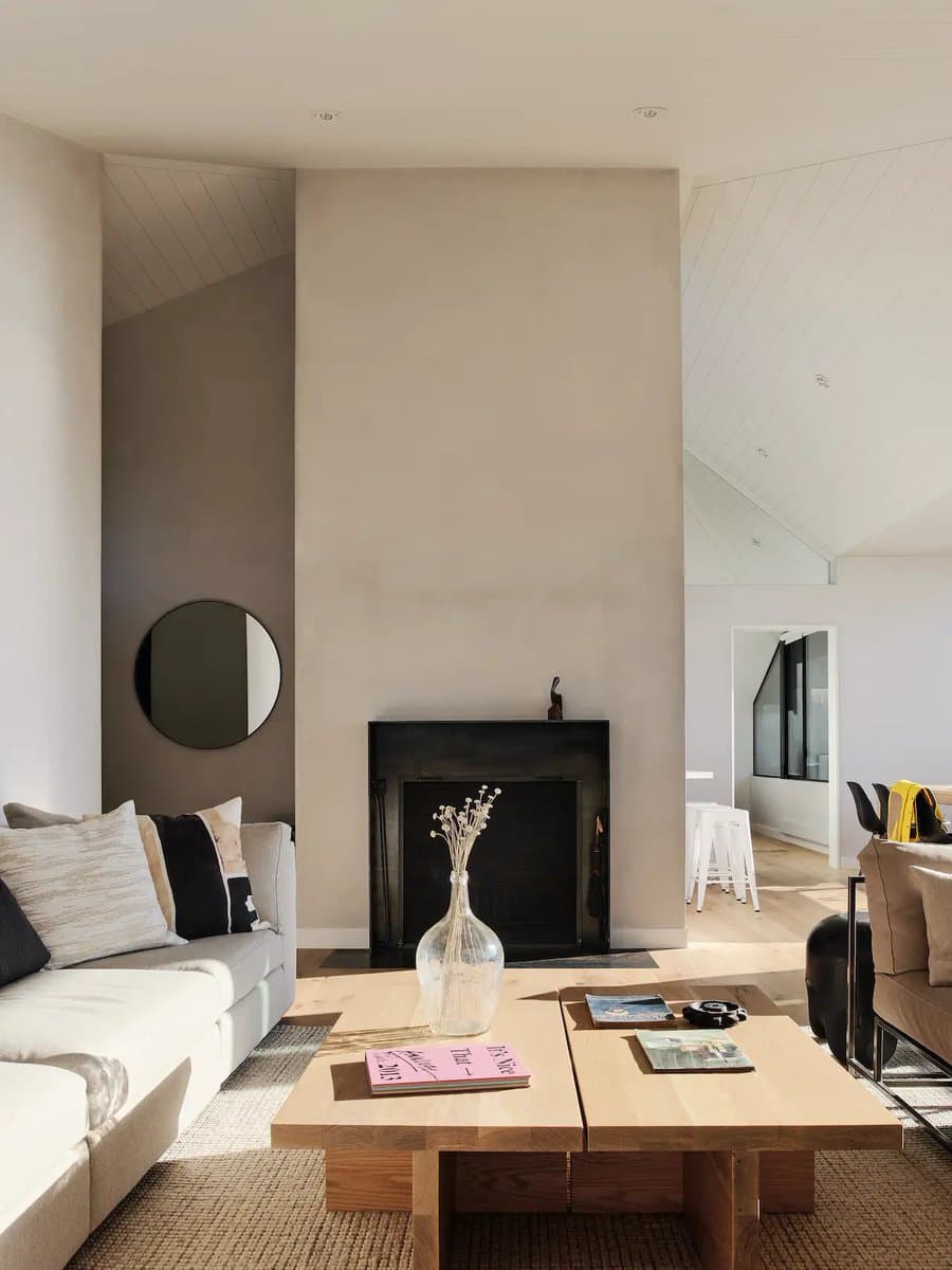 A bright, minimalist living room featuring a large central fireplace, a white sectional sofa, and a low wooden coffee table. The space has high, vaulted ceilings with white paneling and is decorated with neutral tones and simple furnishings.