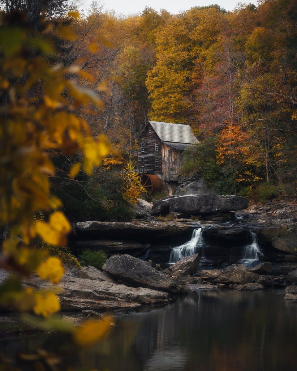 A scenic landscape photograph of a rustic wooden grist mill with a water wheel, nestled among trees with vibrant autumn foliage. A small waterfall cascades over rocks into a calm stream in the foreground, framed by out-of-focus yellow leaves on the left.