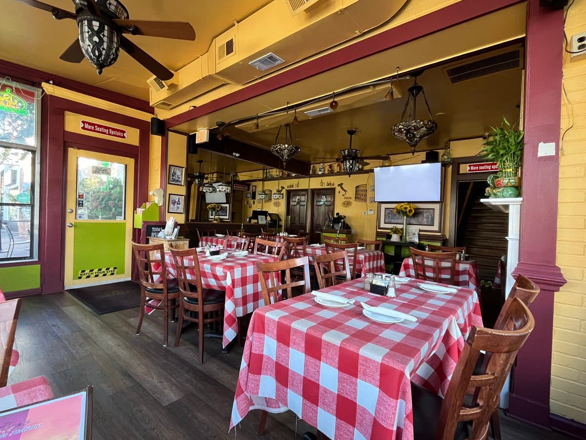 A photograph of an empty restaurant dining room featuring several tables covered in red and white checkered tablecloths, wooden chairs, and hanging light fixtures. The room has yellow walls, a ceiling fan, and a doorway leading to a staircase.