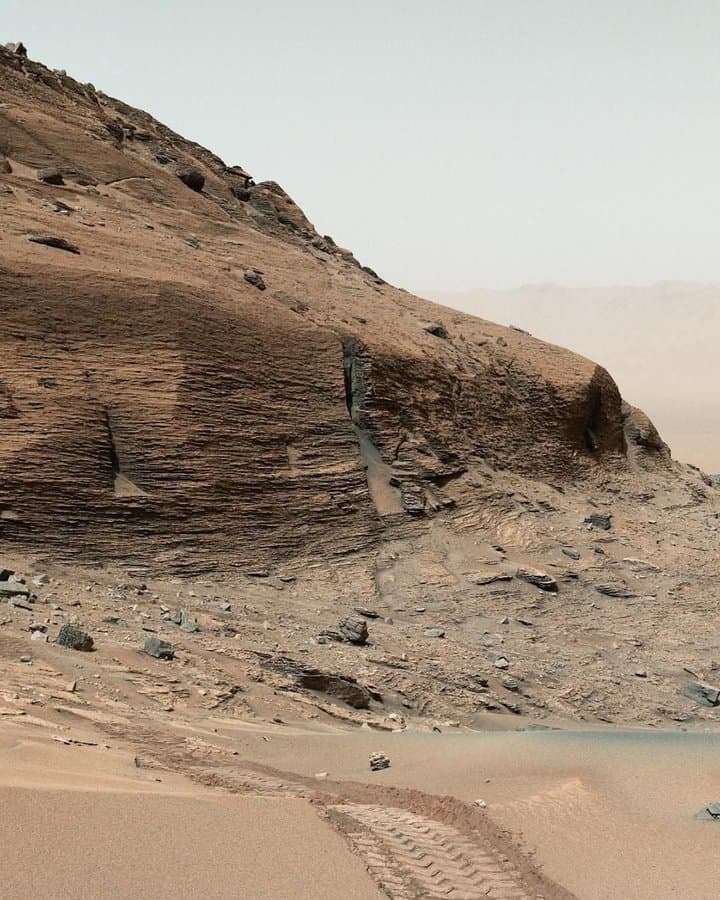 A landscape photograph of the Martian surface showing a steep, layered rock formation with a hazy sky in the background. In the foreground, distinct tire tracks from a rover are visible in the sandy terrain.