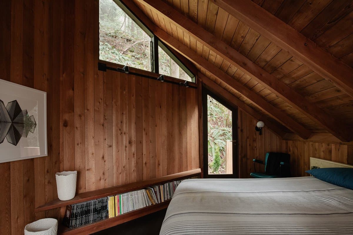 A photograph of a cozy bedroom interior with wood-paneled walls and a sloped ceiling, featuring a bed, a built-in bookshelf, and large windows looking out onto a forest.