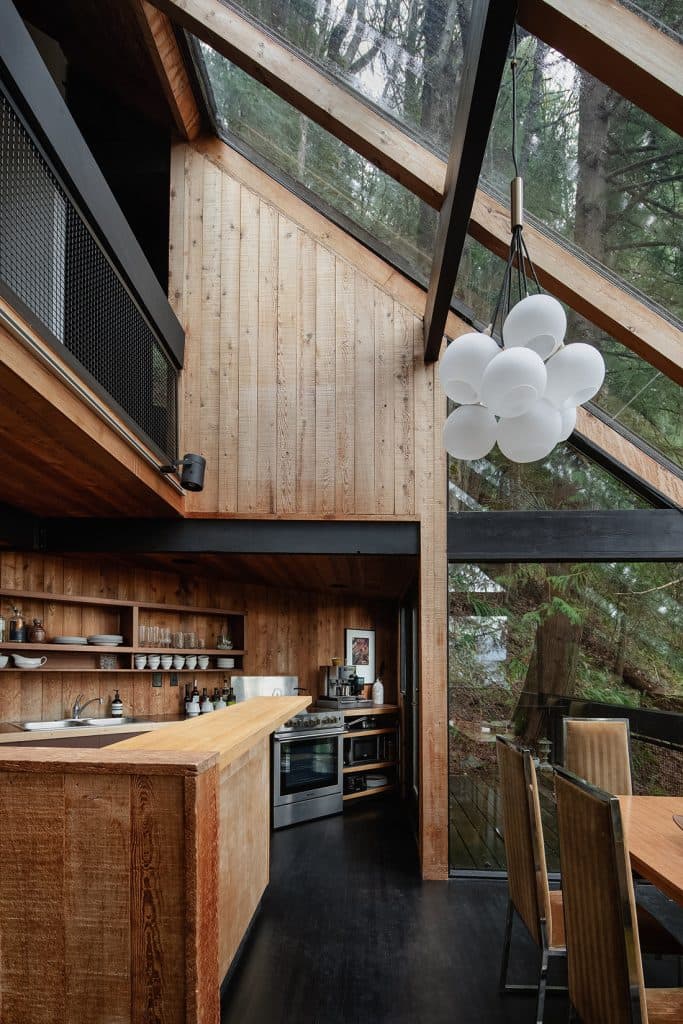 A photograph of a modern tree house interior featuring warm wood paneling, a kitchen with a large wooden island, and a dining area. The space is characterized by a high, sloped ceiling with large glass windows that offer a view of the surrounding forest, and a distinctive cluster of white spherical pendant lights hangs from the ceiling.