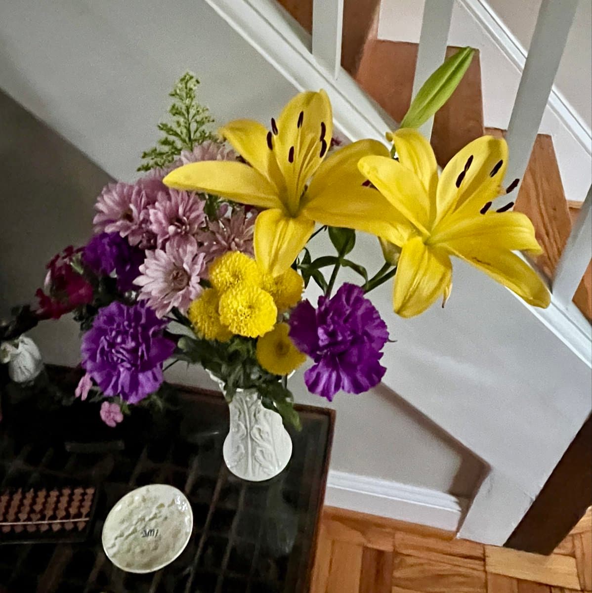 A photograph of a floral arrangement in a white vase, featuring yellow lilies, purple and pink chrysanthemums, and purple carnations, sitting on a dark table near a staircase.