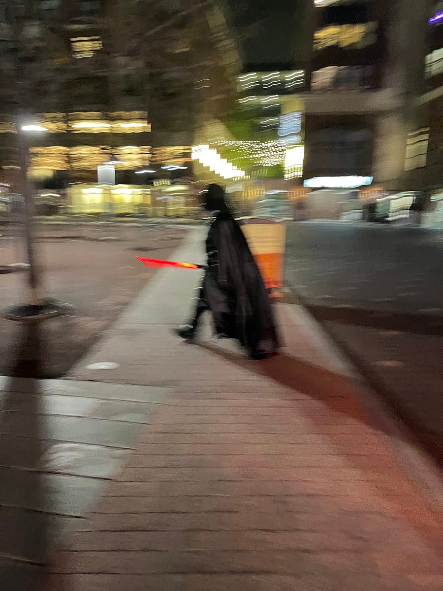 A blurry, low-light photograph of a person walking on a city sidewalk at night. The individual is wearing a long black cape and carrying a glowing red object, possibly a prop sword.