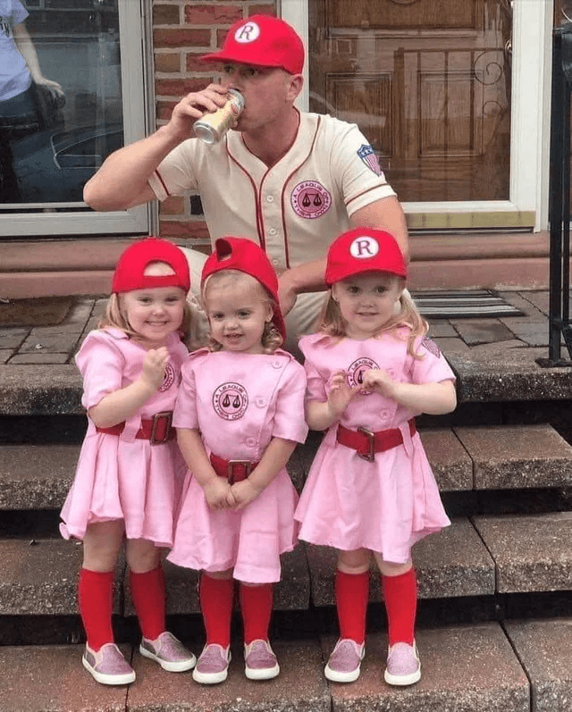 A man and three young girls dressed in vintage baseball uniforms, featuring red caps with the letter 'R' and pink or cream jerseys, posing on a set of outdoor steps.