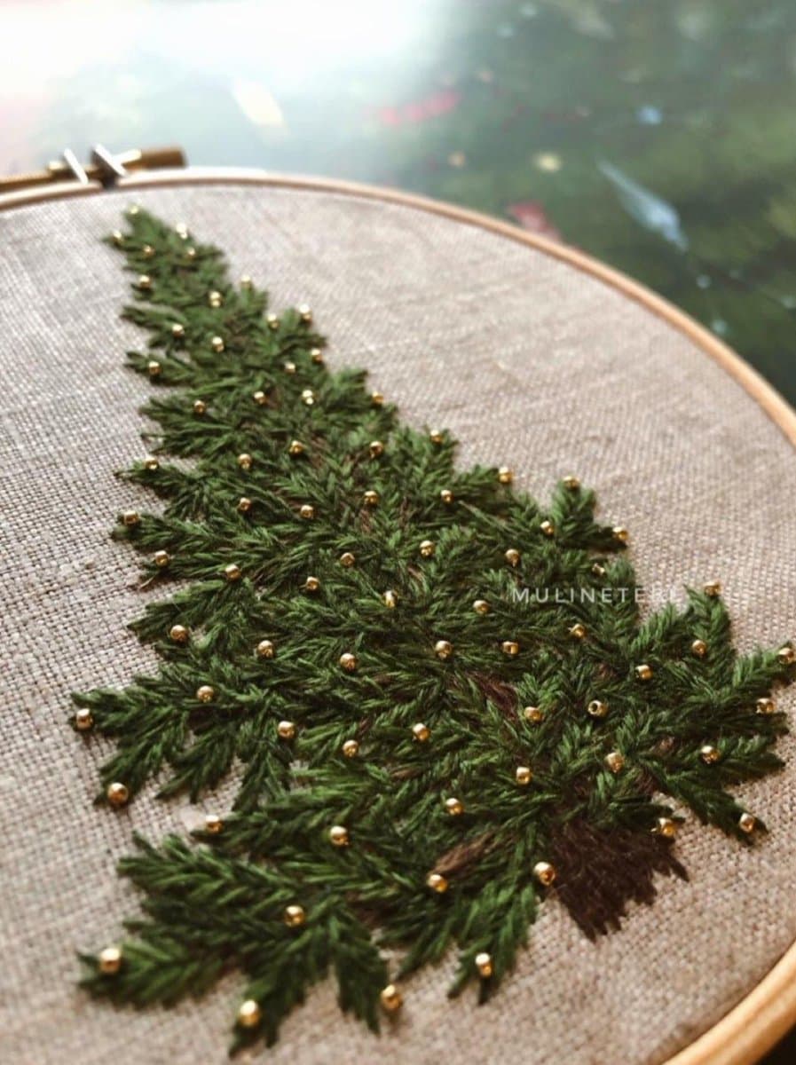 A close-up photograph of a hand-embroidered Christmas tree on a piece of fabric stretched in a wooden hoop. The tree is stitched with green thread and decorated with small gold beads, resembling ornaments.