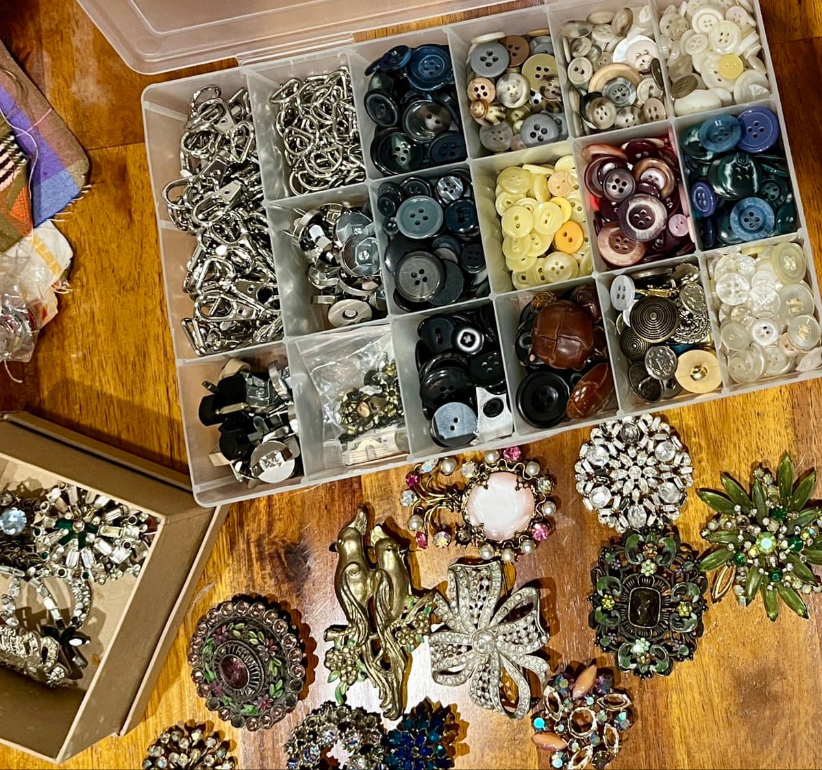 A top-down view of a clear plastic organizer box filled with sorted buttons and metal clasps, sitting on a wooden surface next to an assortment of decorative brooches and jewelry pieces.