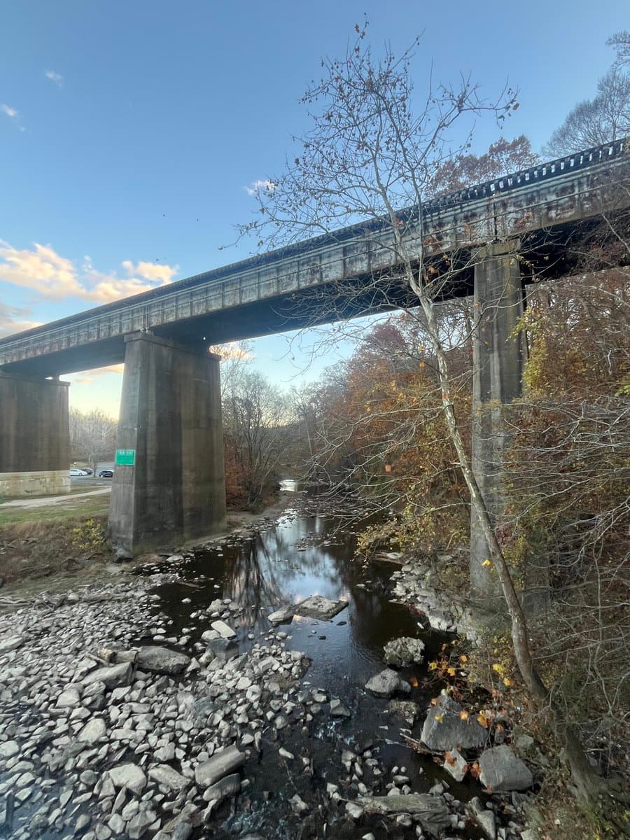 A photograph showing a concrete bridge spanning a rocky stream under a blue sky with autumn foliage.