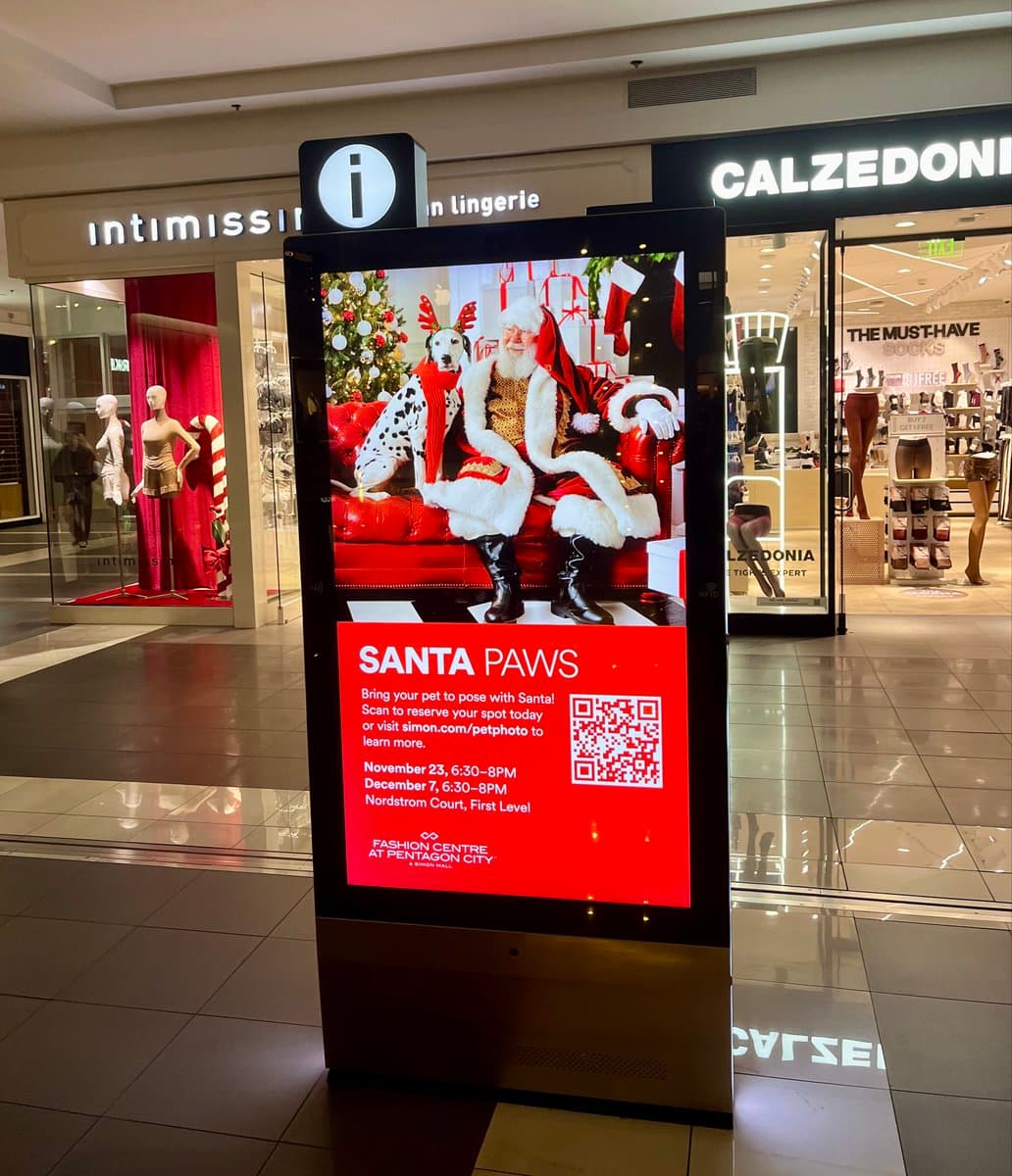 A digital advertising kiosk in a shopping mall displays a holiday promotion for a pet photo-op with Santa. The screen shows a photo of Santa Claus sitting on a red couch with a Dalmatian dog wearing a red scarf, set against a festive background with a Christmas tree and gifts.