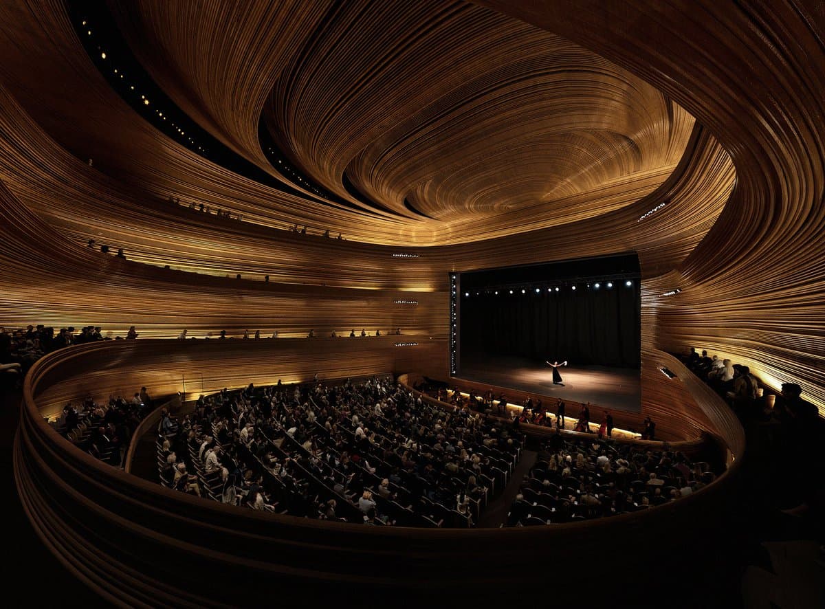 A wide-angle photograph of a modern concert hall featuring a sweeping, curved wooden ceiling and tiered seating filled with an audience. A single performer stands on the illuminated stage at the far end of the hall.
