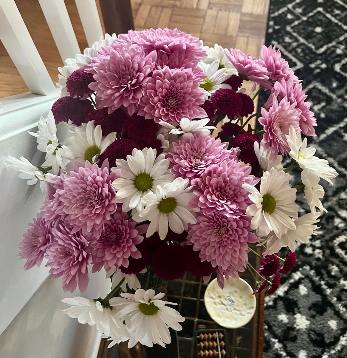 A close-up photograph of a large bouquet of flowers, featuring pink and white daisies and deep purple chrysanthemums, resting on a white surface next to a patterned rug. The image is a standard photo, not a text-bearing graphic.