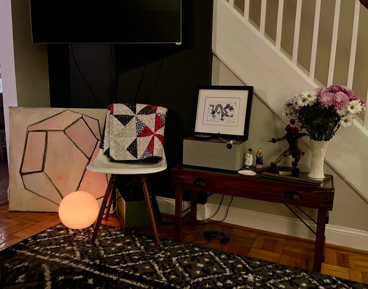 A photograph of a home interior featuring a white chair with a red, black, and white quilted pillow, a glowing spherical floor lamp, and a wooden console table holding a framed picture, a speaker, and a vase of flowers.