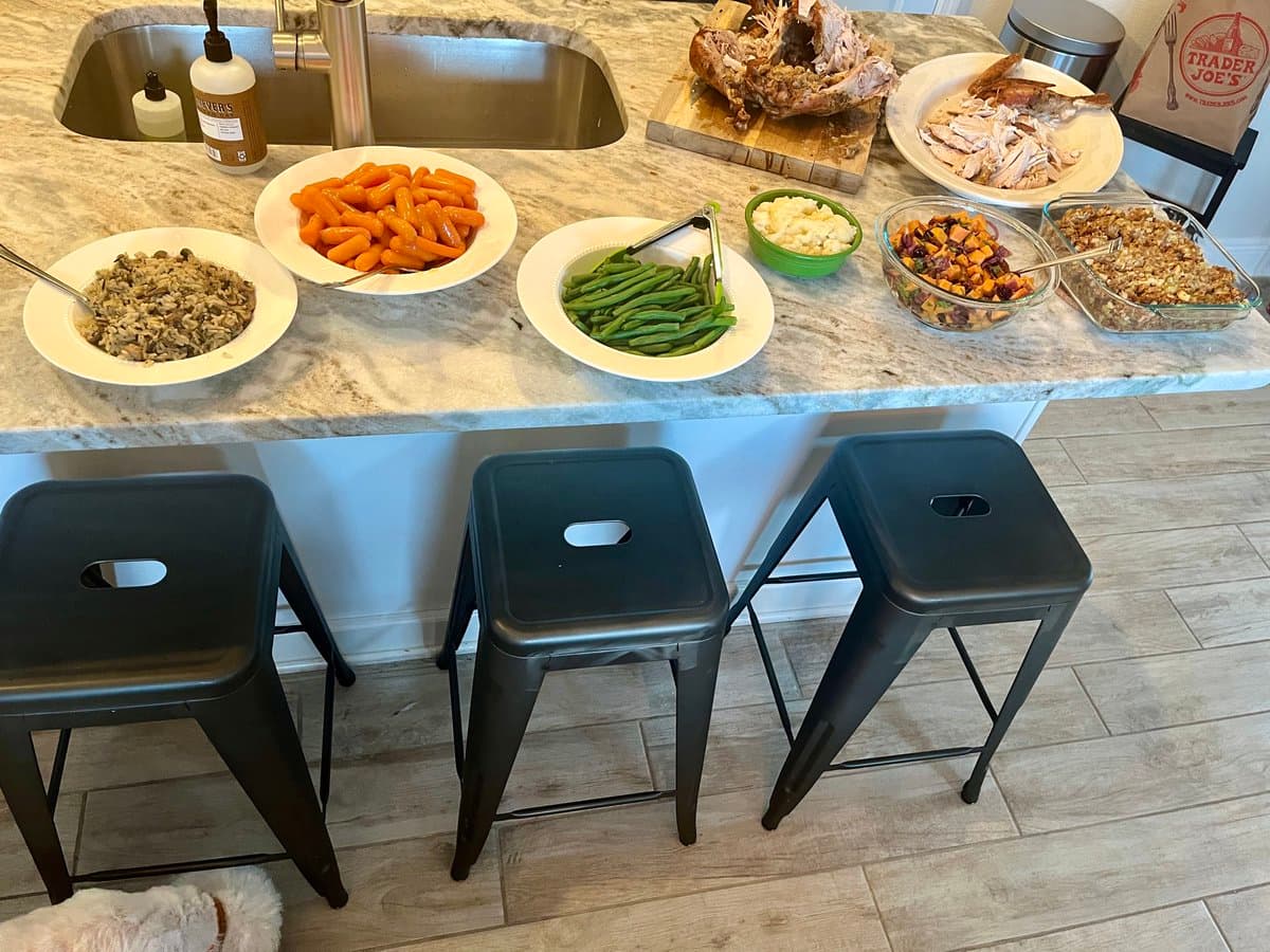 A kitchen island is set with a Thanksgiving-style meal, including a carved turkey, mashed potatoes, green beans, sweet potatoes, stuffing, and a salad, with three black metal stools in the foreground. A Trader Joe's bag is visible in the background.