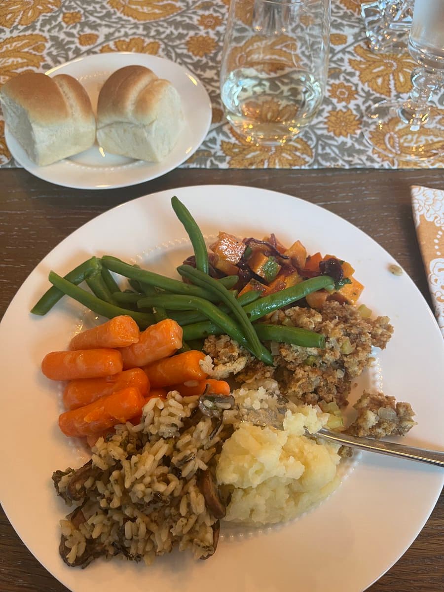 A plate of food featuring green beans, baby carrots, wild rice, mashed potatoes, stuffing, and a vegetable medley, accompanied by a side plate of dinner rolls and a glass of water on a patterned tablecloth.