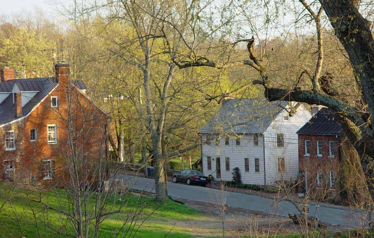 A scenic photograph of a quiet, tree-lined residential street in a small village, featuring a red brick house on the left and a white clapboard house on the right, with a parked car and bare trees in the foreground.