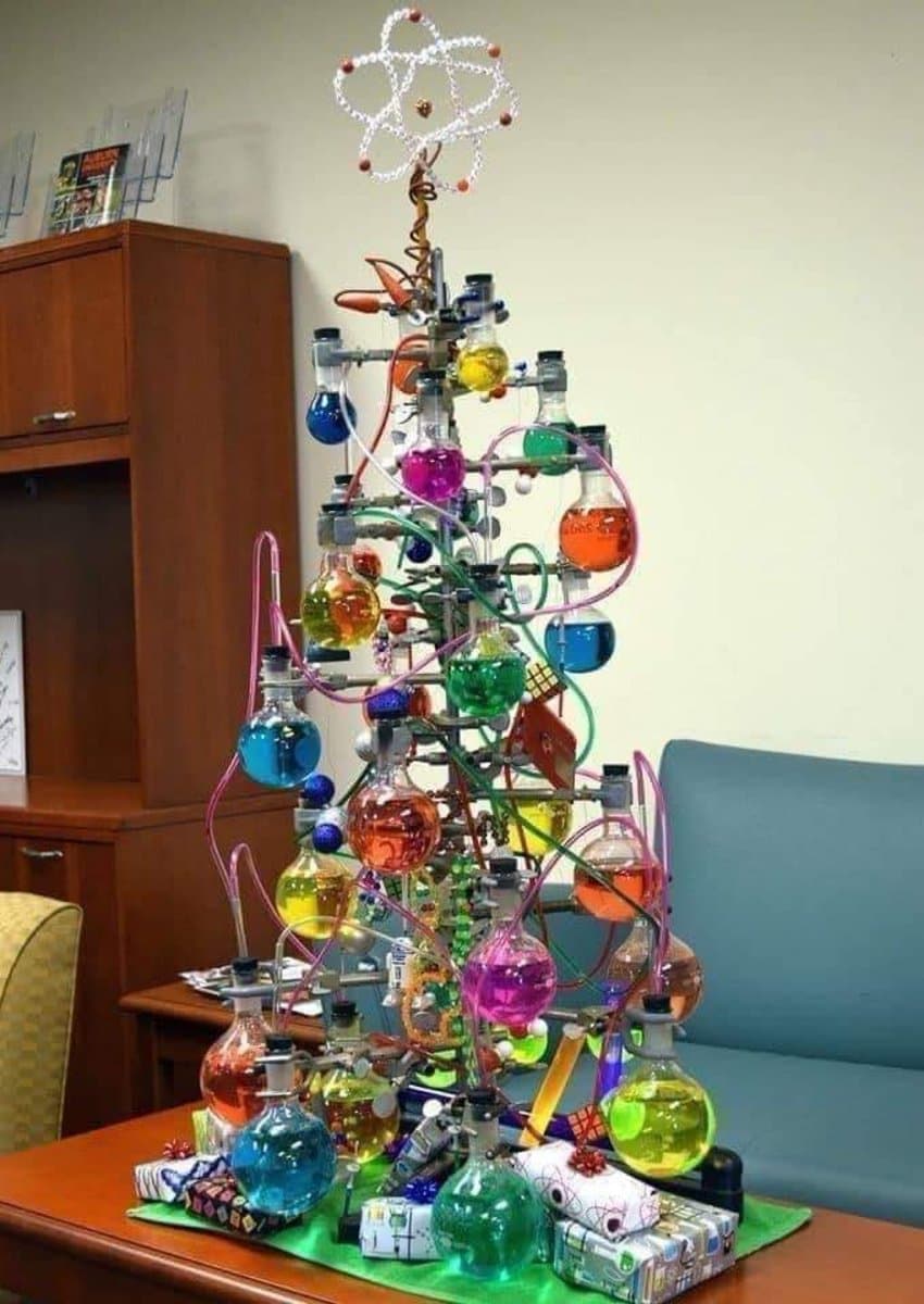 A creative Christmas tree made from laboratory glassware, including flasks and beakers filled with colorful liquids, topped with a wire atom model. The tree is decorated with small wrapped gifts and stands on a table in an office setting.