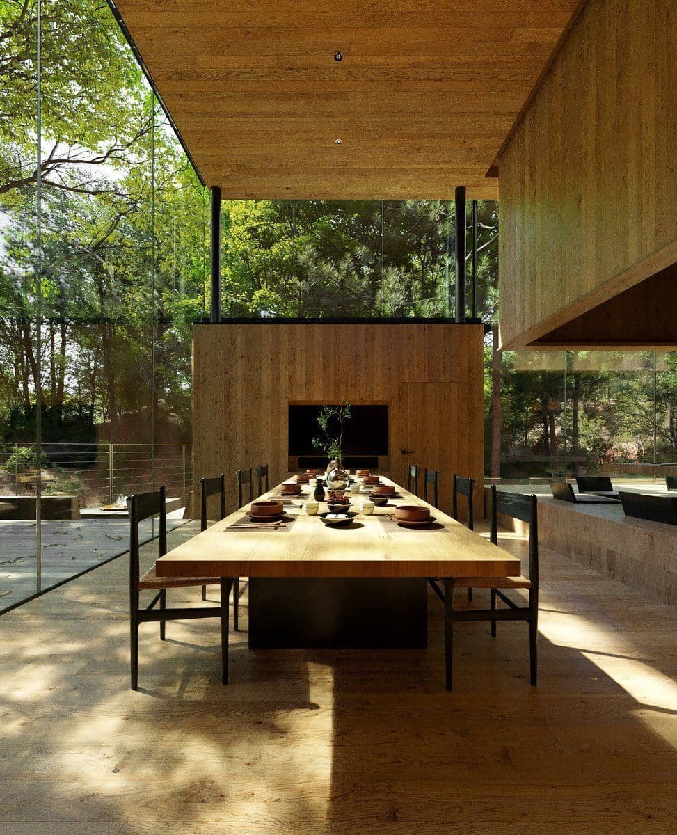 A photograph of a modern dining room featuring a long wooden table set for a meal, surrounded by black chairs. The space is enclosed by floor-to-ceiling glass walls that look out onto a dense forest, with a wooden ceiling and a built-in fireplace in the background.