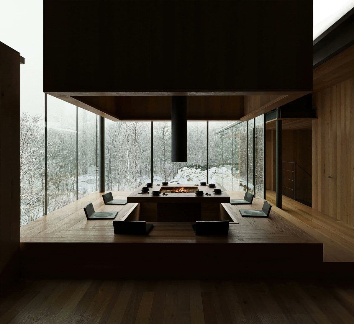 A modern, minimalist dining room with a long wooden table and chairs, set on a raised platform. Floor-to-ceiling glass windows offer a panoramic view of a snow-covered forest, with a central fireplace built into the table.