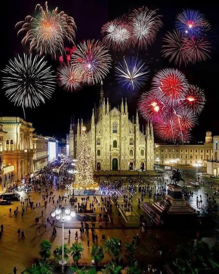 A nighttime photograph of the Piazza del Duomo in Milan, featuring the illuminated Milan Cathedral and a large Christmas tree in the foreground. Colorful fireworks burst in the dark sky above the cathedral, while crowds of people gather in the square below.