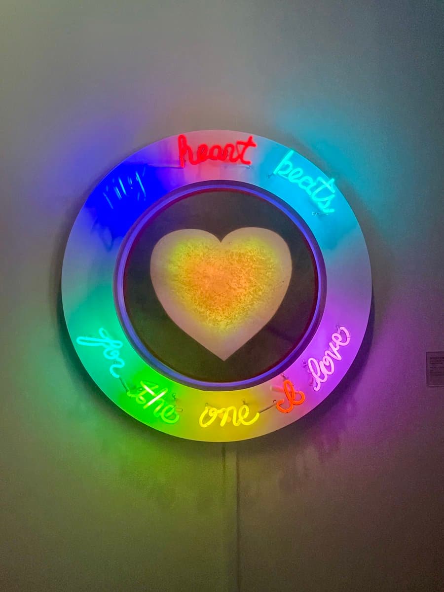 A circular neon light art installation mounted on a wall, featuring a central yellow heart surrounded by a rainbow-colored ring. The ring contains the cursive text 'heart beats for the one & love' in various neon colors.