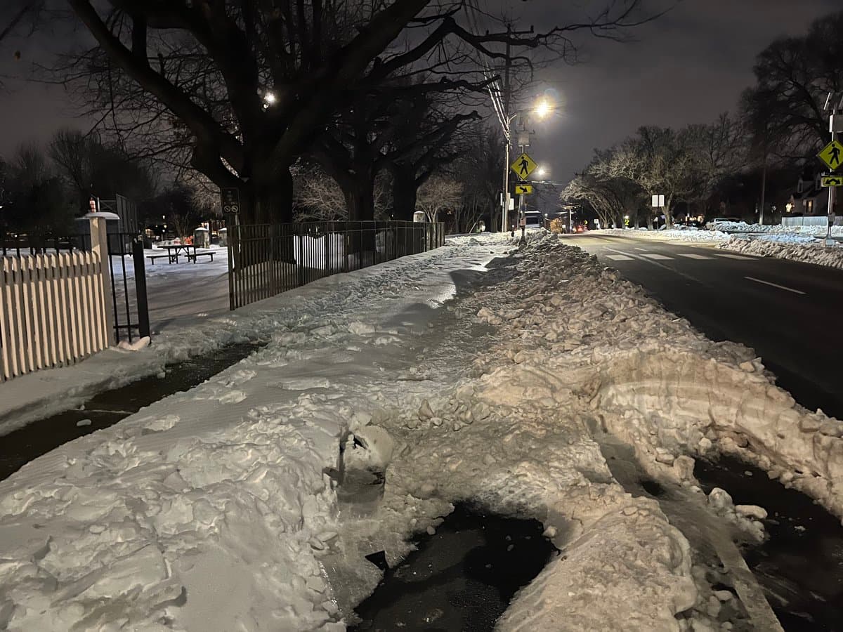 A nighttime photograph of a snow-covered sidewalk and street, with large piles of plowed snow lining the road. Bare trees, a fence, and streetlights are visible in the background, creating a cold, wintry scene.