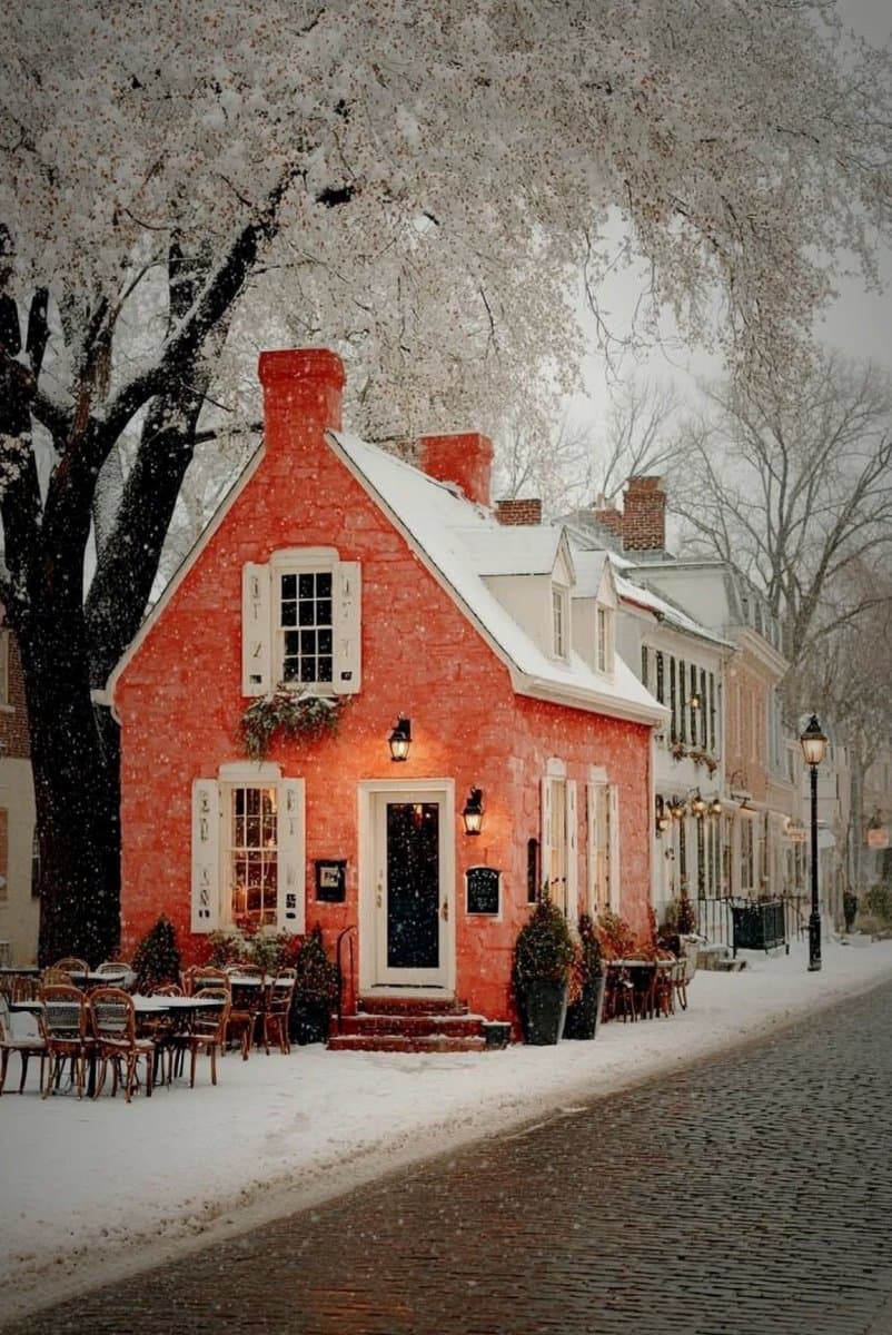A red brick building with white trim and a snow-covered roof sits on a cobblestone street during a snowfall, with outdoor dining tables and chairs set up in the snow.