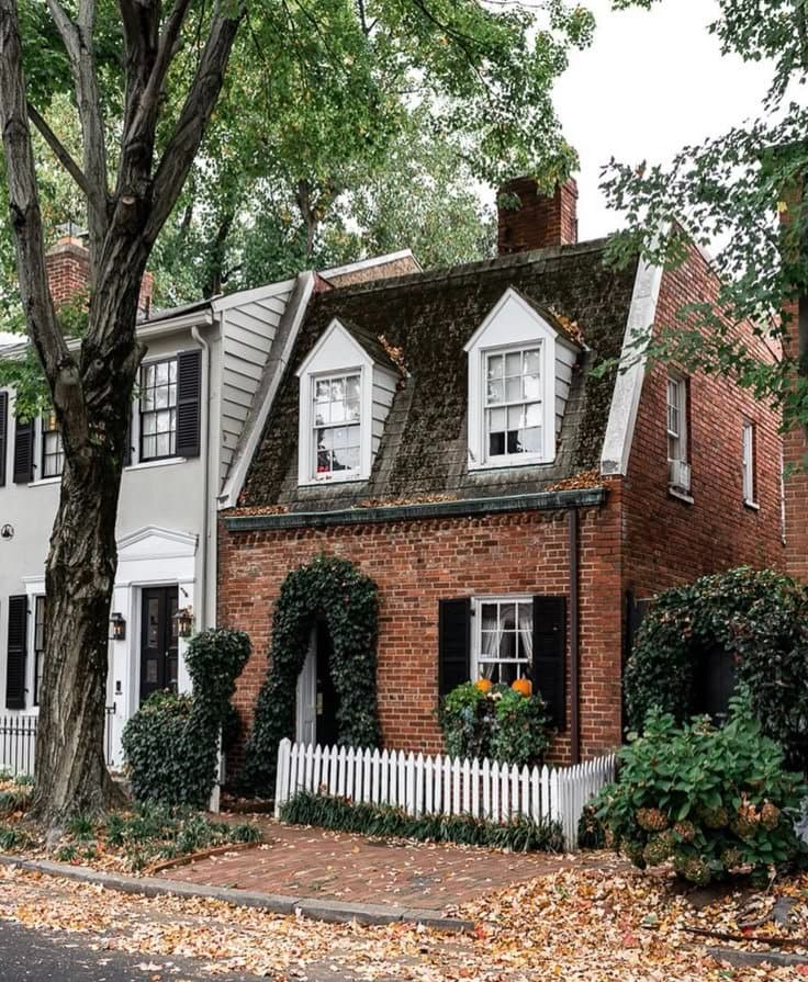 A photograph of a charming, two-story brick house with a mossy roof and white picket fence, surrounded by trees and autumn leaves.