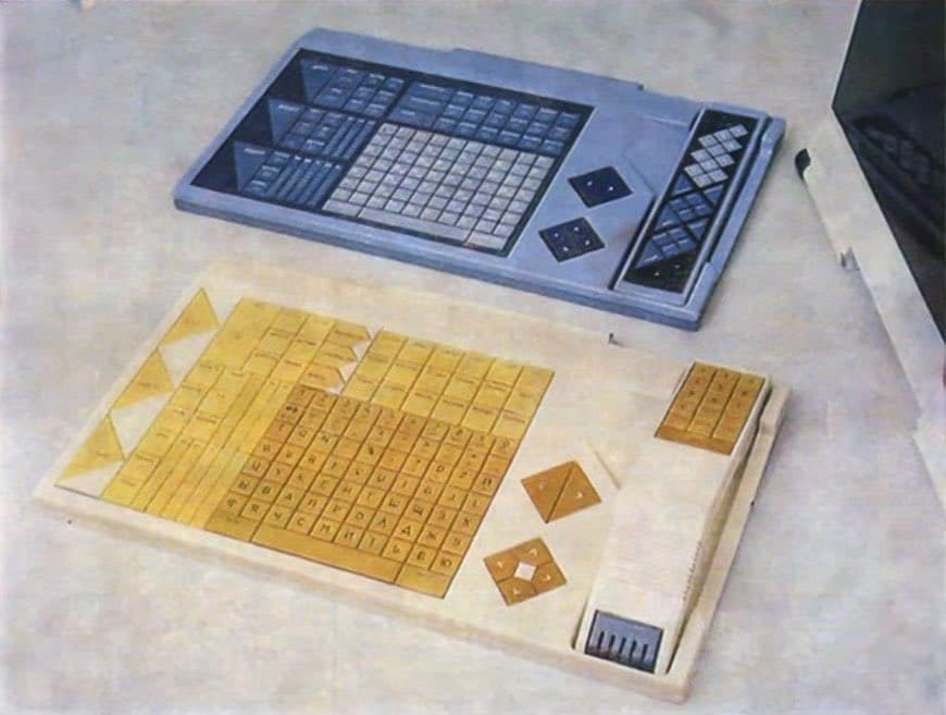 A photograph showing a physical prototype of the SPHINX computer keyboard, featuring a grey plastic model and a wooden mockup with yellow keycaps, resting on a light-colored surface.