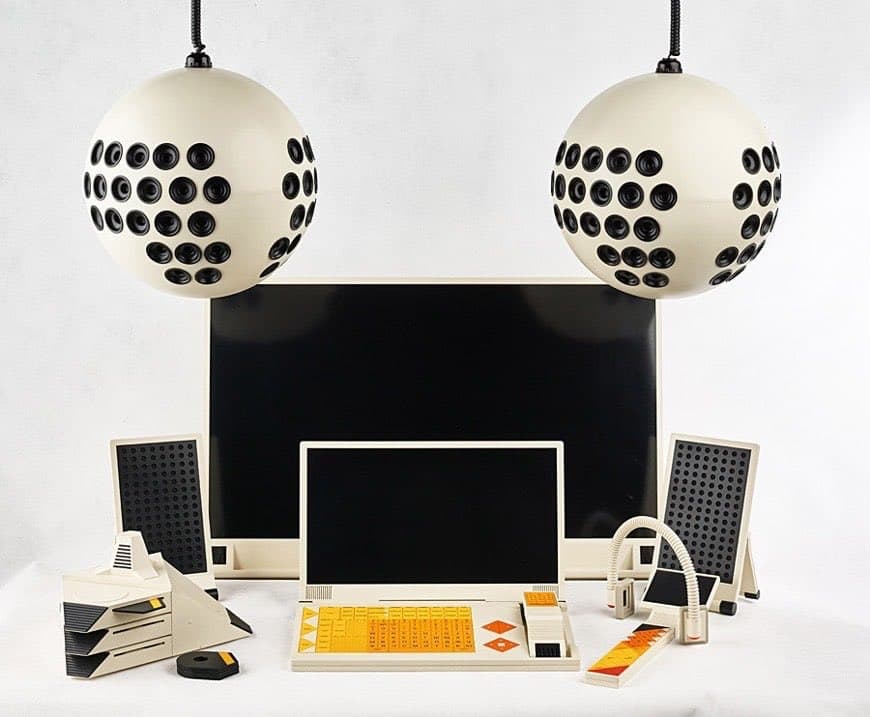 A photograph of a retro-futuristic computer setup featuring a central monitor, a keyboard with yellow keys, and two large spherical speakers suspended from above. The design is minimalist and geometric, with a cream and black color scheme.