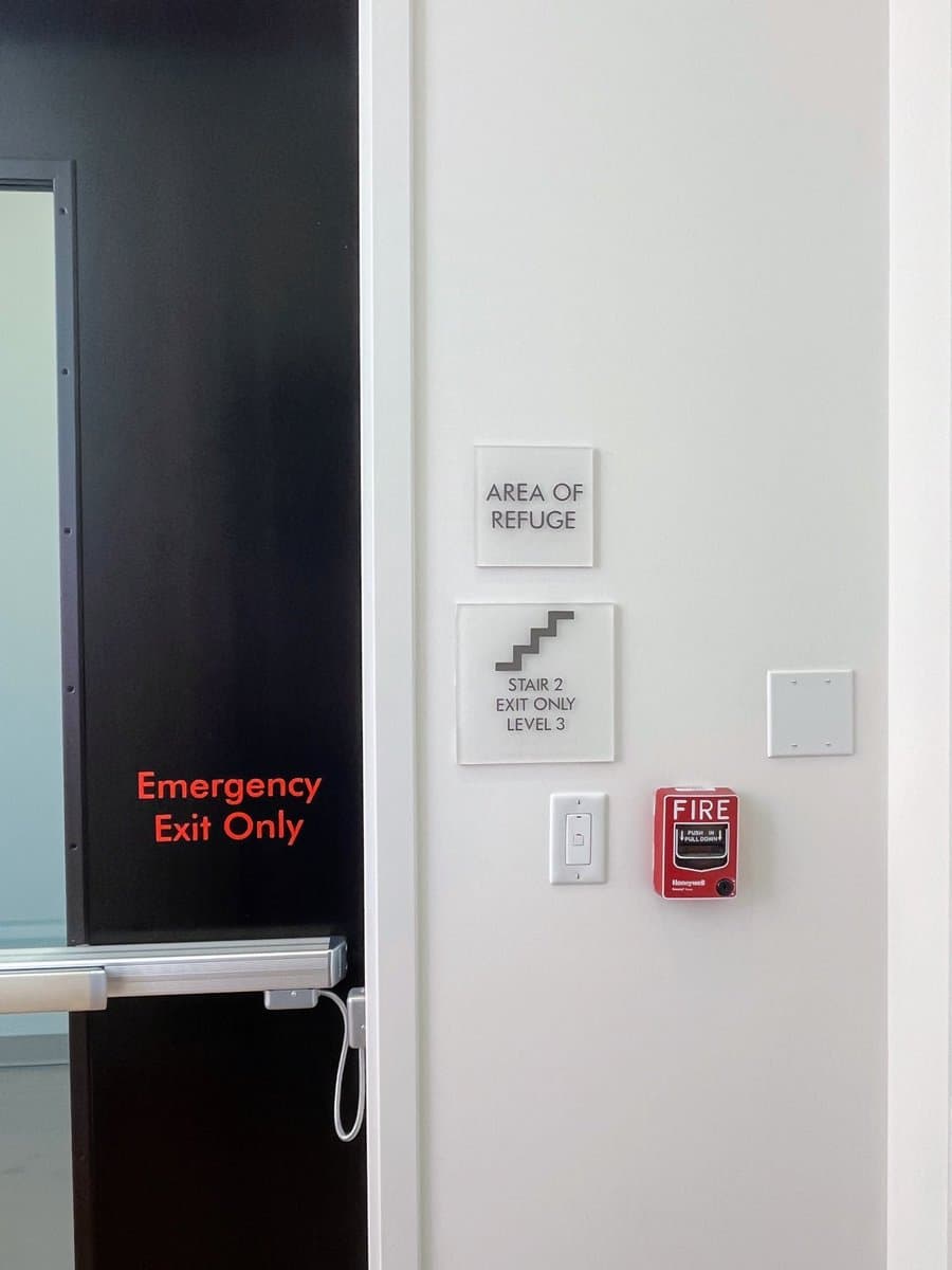 A photo of a black emergency exit door next to a white wall with safety signage and a fire alarm pull station. The signs indicate an 'Area of Refuge' and 'Stair 2 Exit Only Level 3'.
