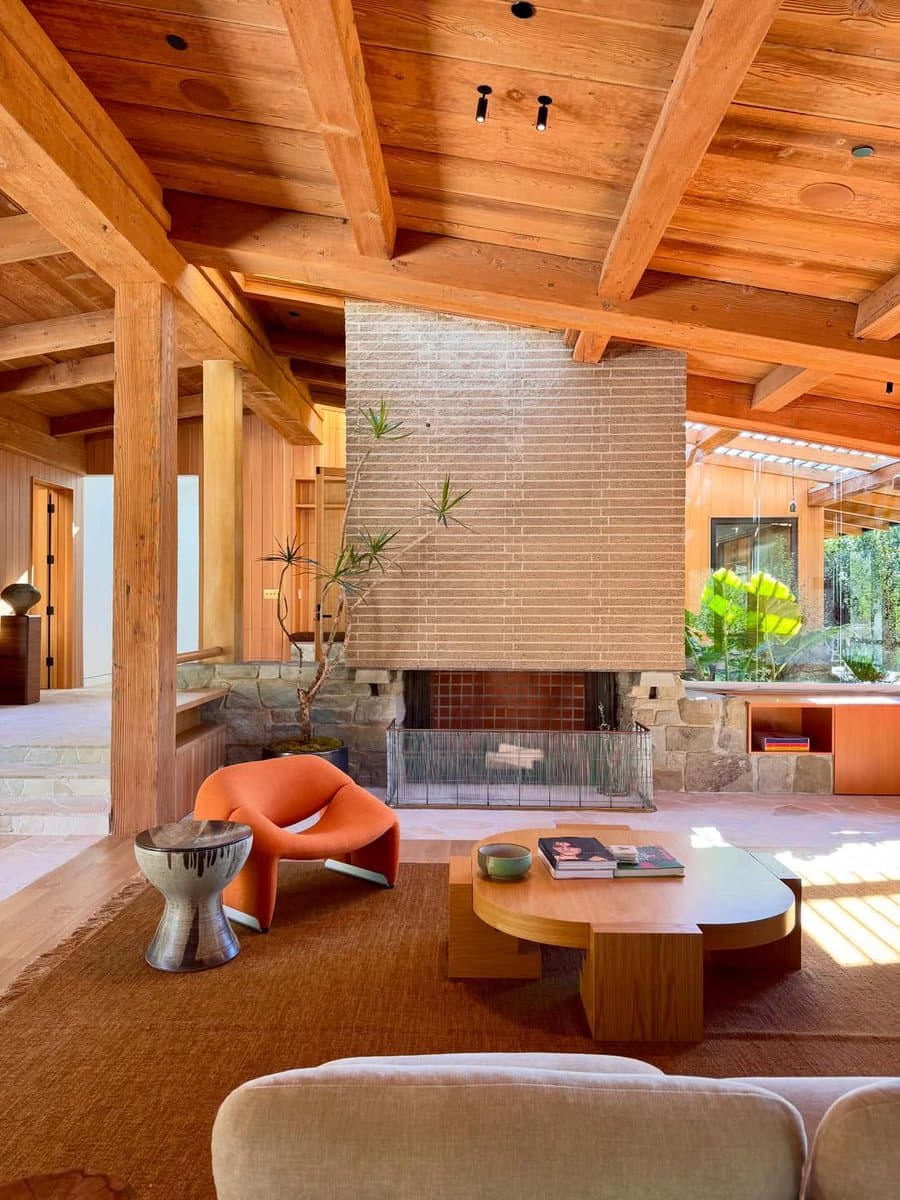 A photograph of a mid-century modern living room featuring exposed wooden ceiling beams, a large brick fireplace, and a low wooden coffee table. The space is furnished with an orange lounge chair, a metallic side table, and a beige sofa, set on a woven rug.