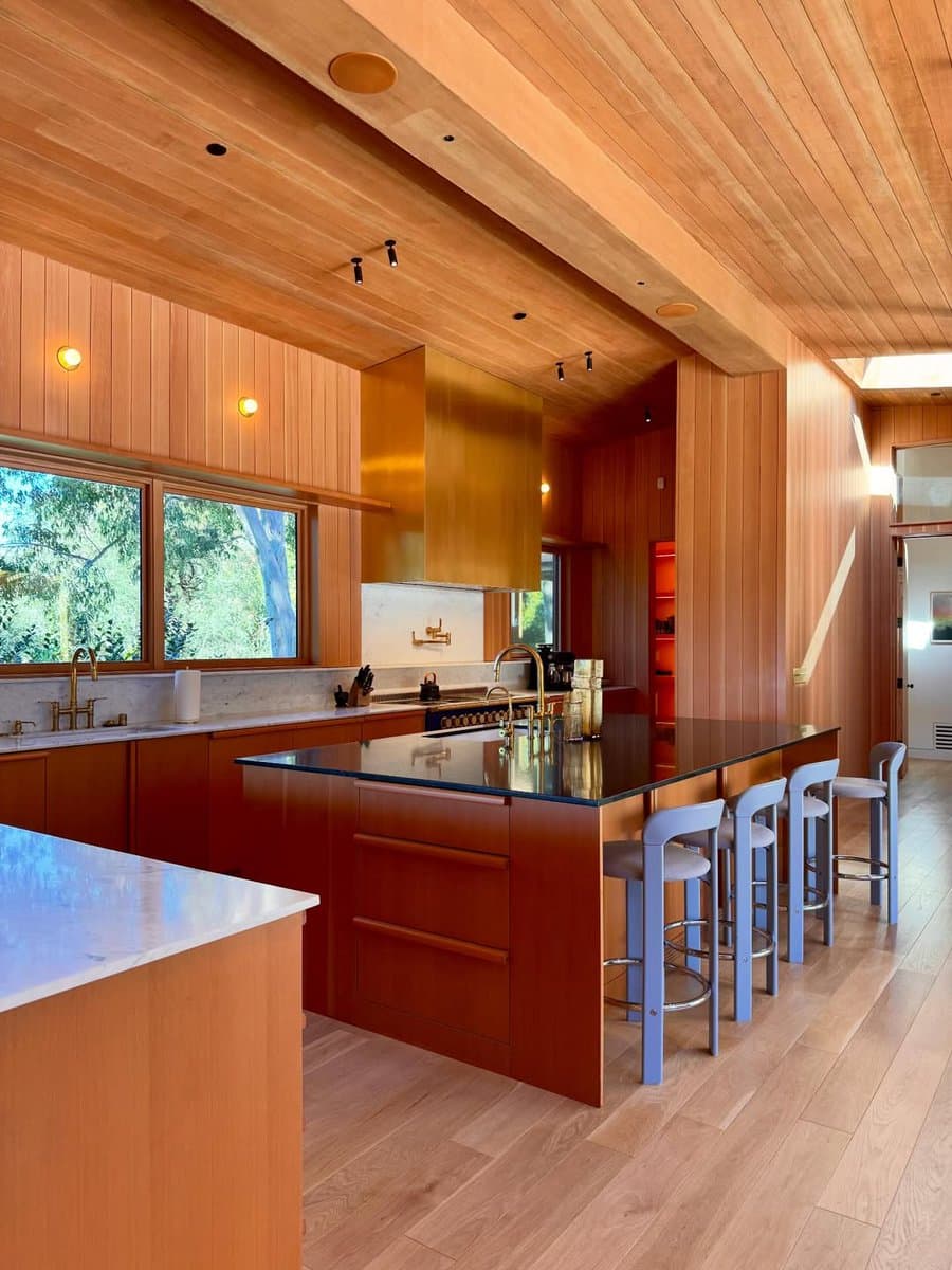A photograph of a mid-century modern kitchen featuring extensive wood paneling on the walls and ceiling, a large central island with a dark countertop, and a gold range hood. Four light blue bar stools are lined up at the island, and large windows on the left look out onto greenery.