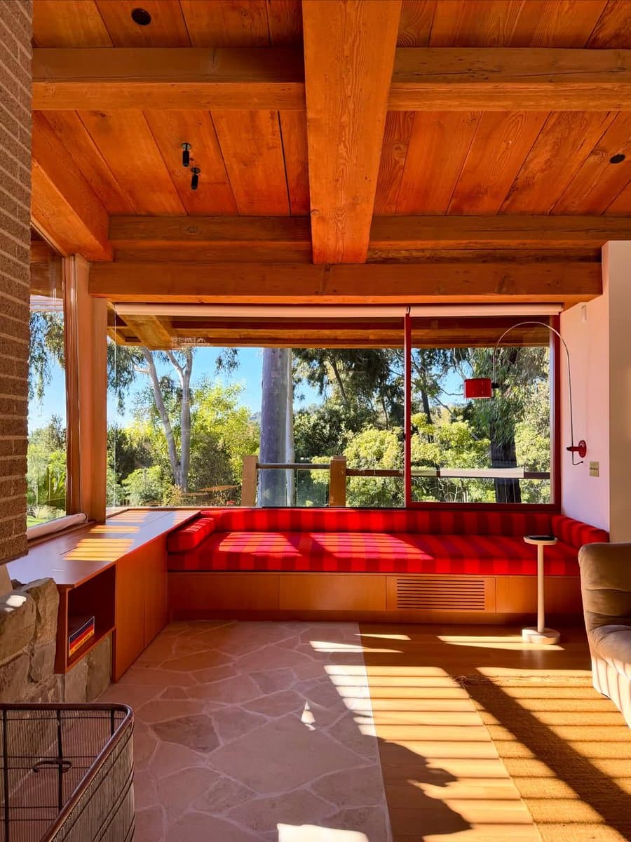 An interior photograph of a mid-century modern living room featuring a built-in red and orange striped bench seat, a wooden ceiling with exposed beams, and large windows looking out onto a wooded landscape.