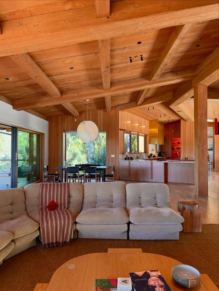 A photograph of a spacious, wood-paneled interior featuring a mid-century modern design with exposed timber beams, a large sectional sofa, and an open-plan kitchen and dining area.