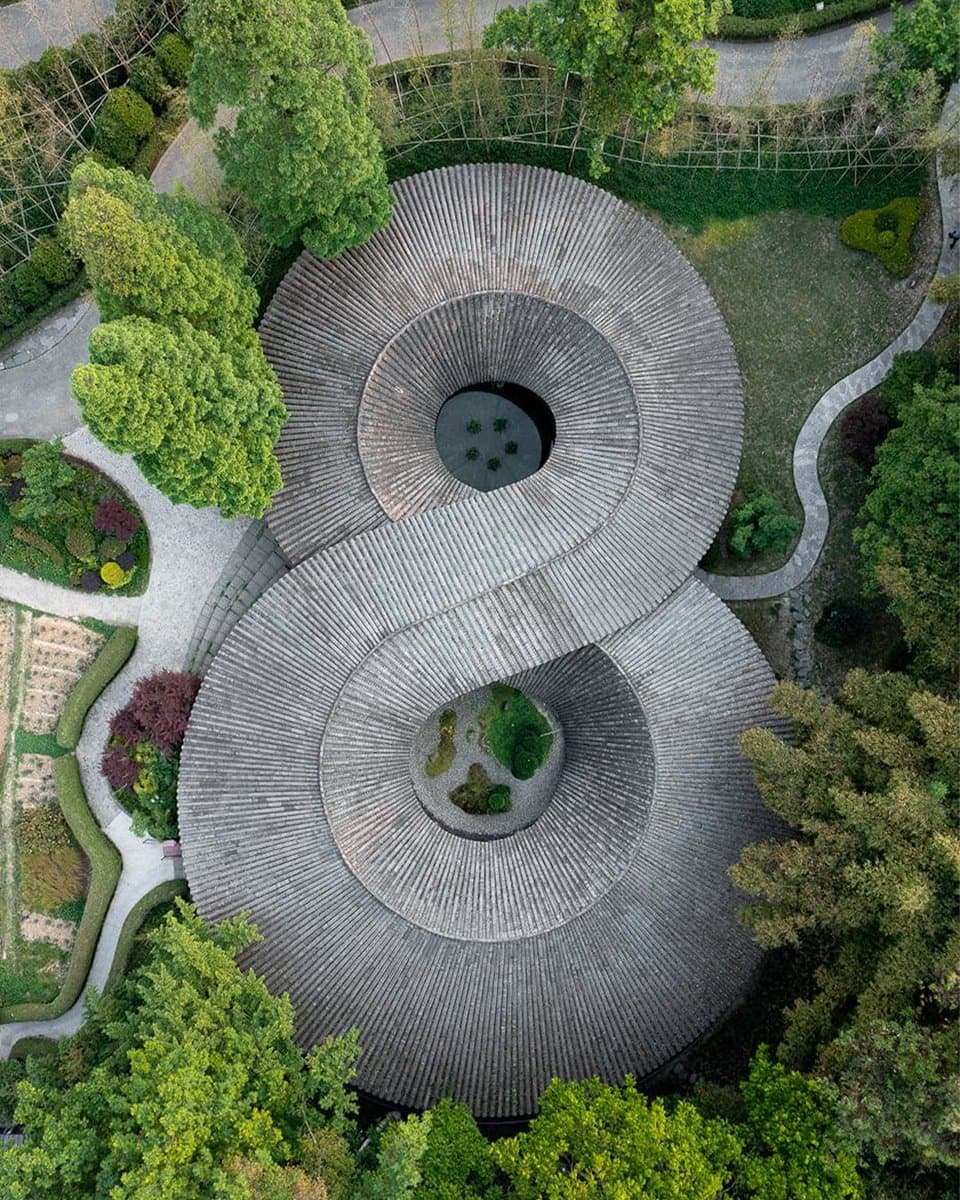 An aerial photograph of a large, circular building with a distinctive spiral roof design, surrounded by lush green trees and landscaped gardens. The structure features two central open courtyards and is situated within a park-like setting with winding pathways.
