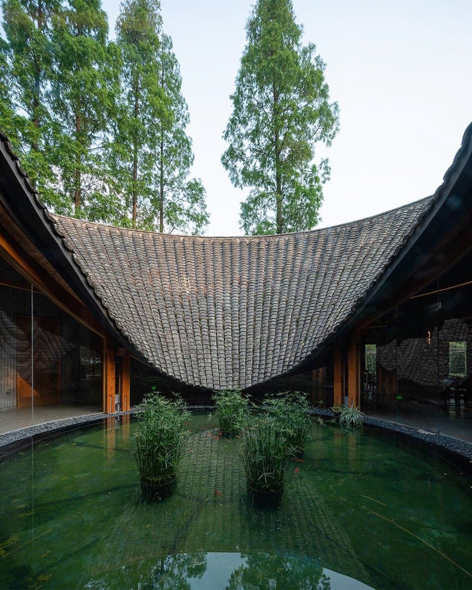 A photograph of a modern architectural structure featuring a large, curved, tiled roof that arches over a central courtyard. The courtyard contains a shallow pool of green water with several clumps of bamboo growing in it, surrounded by glass walls and tall trees in the background.