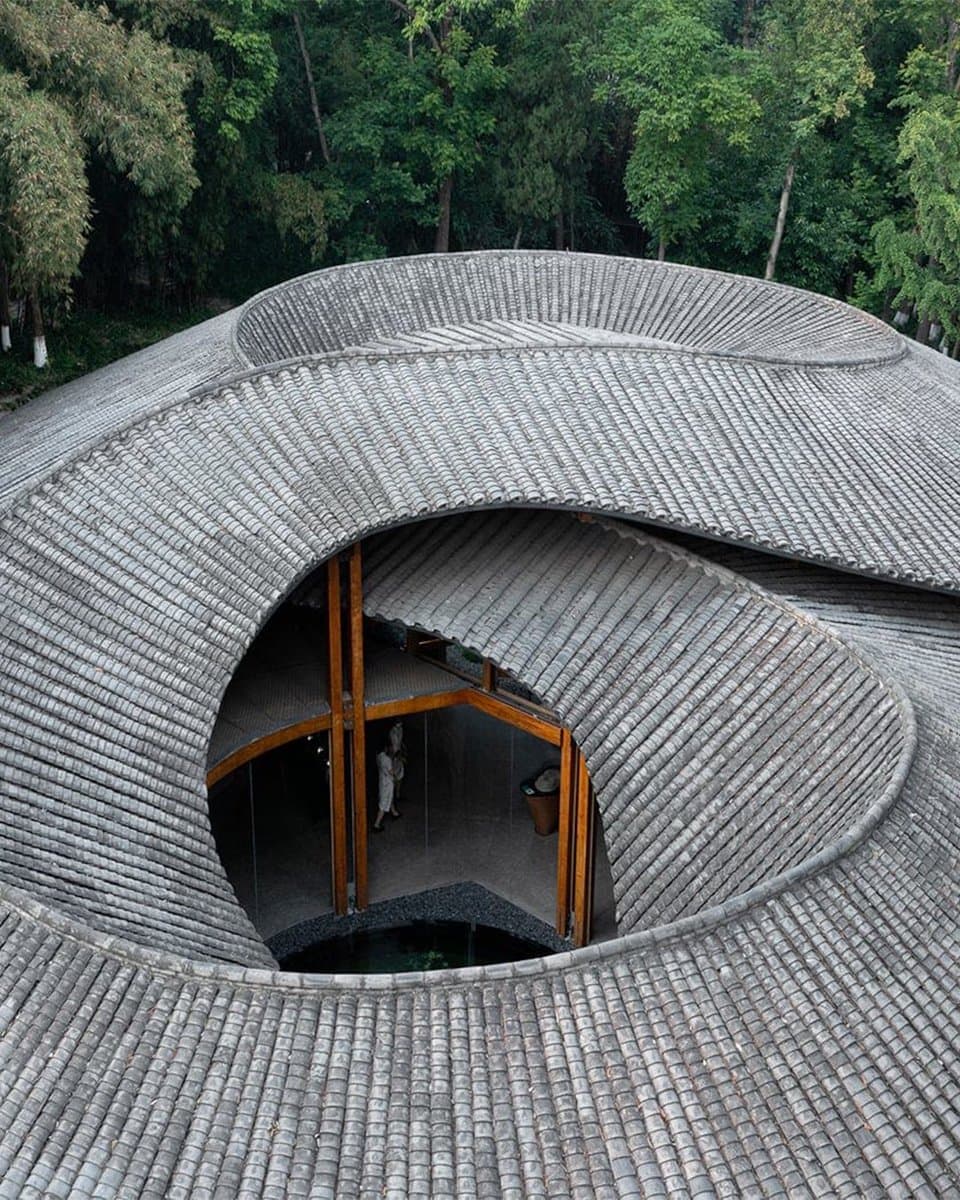 An aerial view of a circular, tunnel-like building with a curved, tiled roof that features a large central opening. The structure is surrounded by a dense forest of green trees, and a person can be seen standing inside the open interior space.