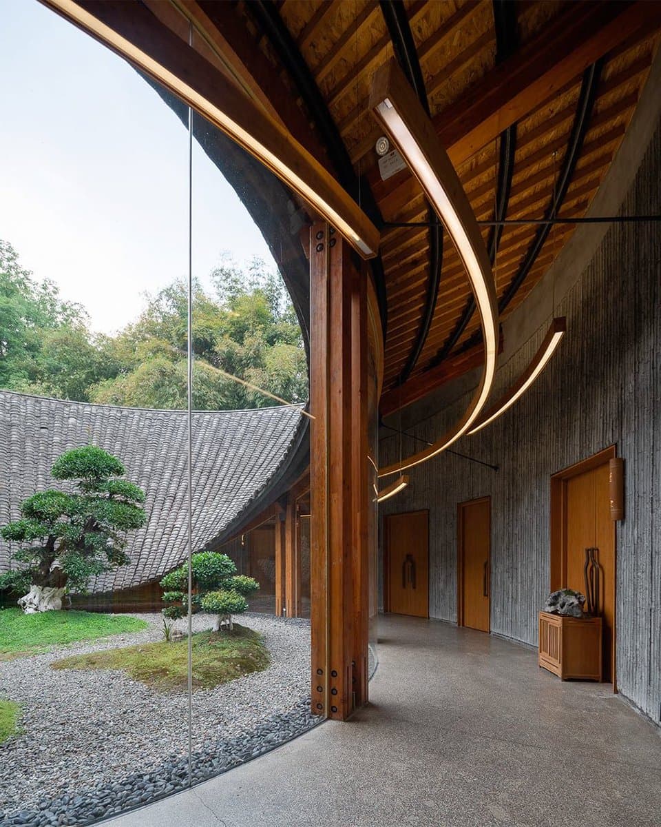 An architectural photograph showing the interior of a building with a curved wooden ceiling and bamboo-style walls, looking out through a large glass wall onto a traditional Japanese-style garden with a curved tiled roof and manicured trees.
