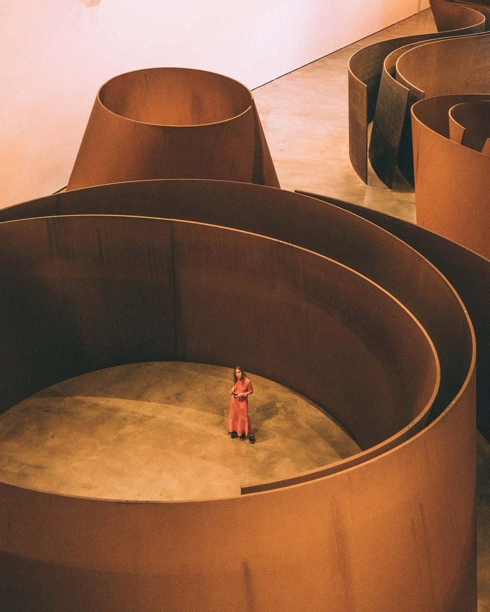 A high-angle photograph of a person in a long red dress standing inside a massive, curved, rust-colored steel sculpture in a gallery space. The image shows the monumental scale of the artwork, which consists of several large, folded metal forms.